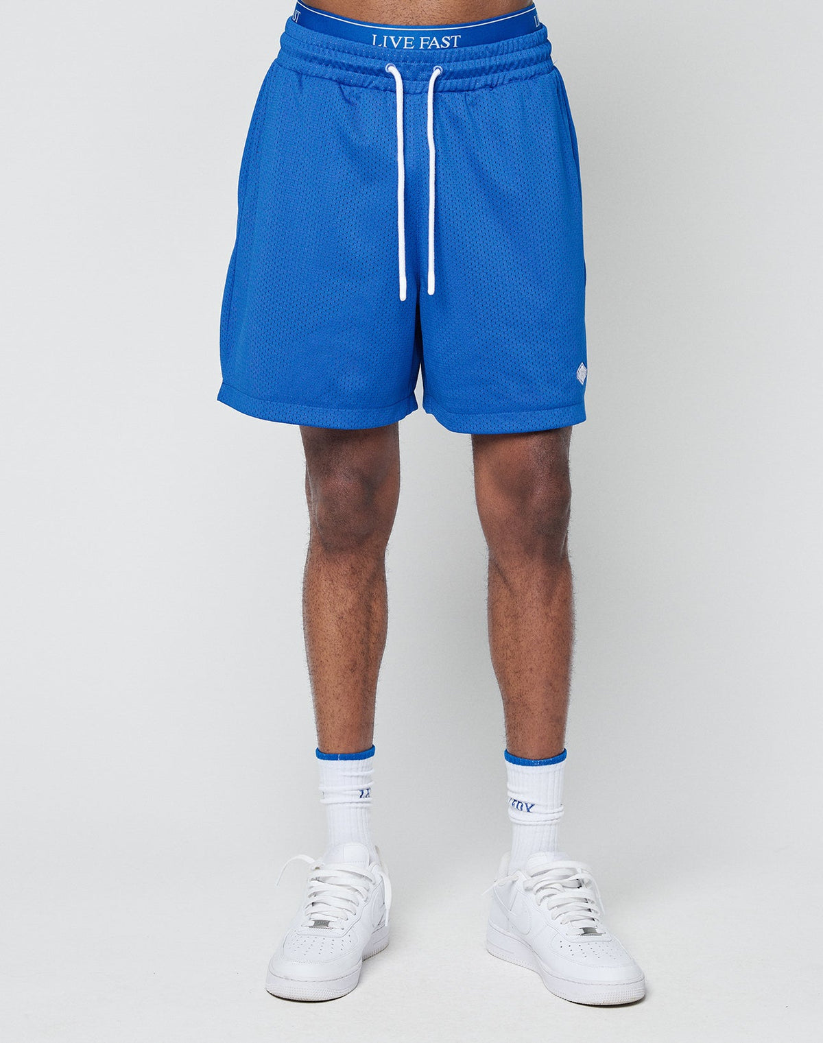 A person wears blue Basic Mesh Shorts by LIVE FAST DIE YOUNG, featuring an elastic waistband, paired with white socks and sneakers, standing against a plain light gray background.
