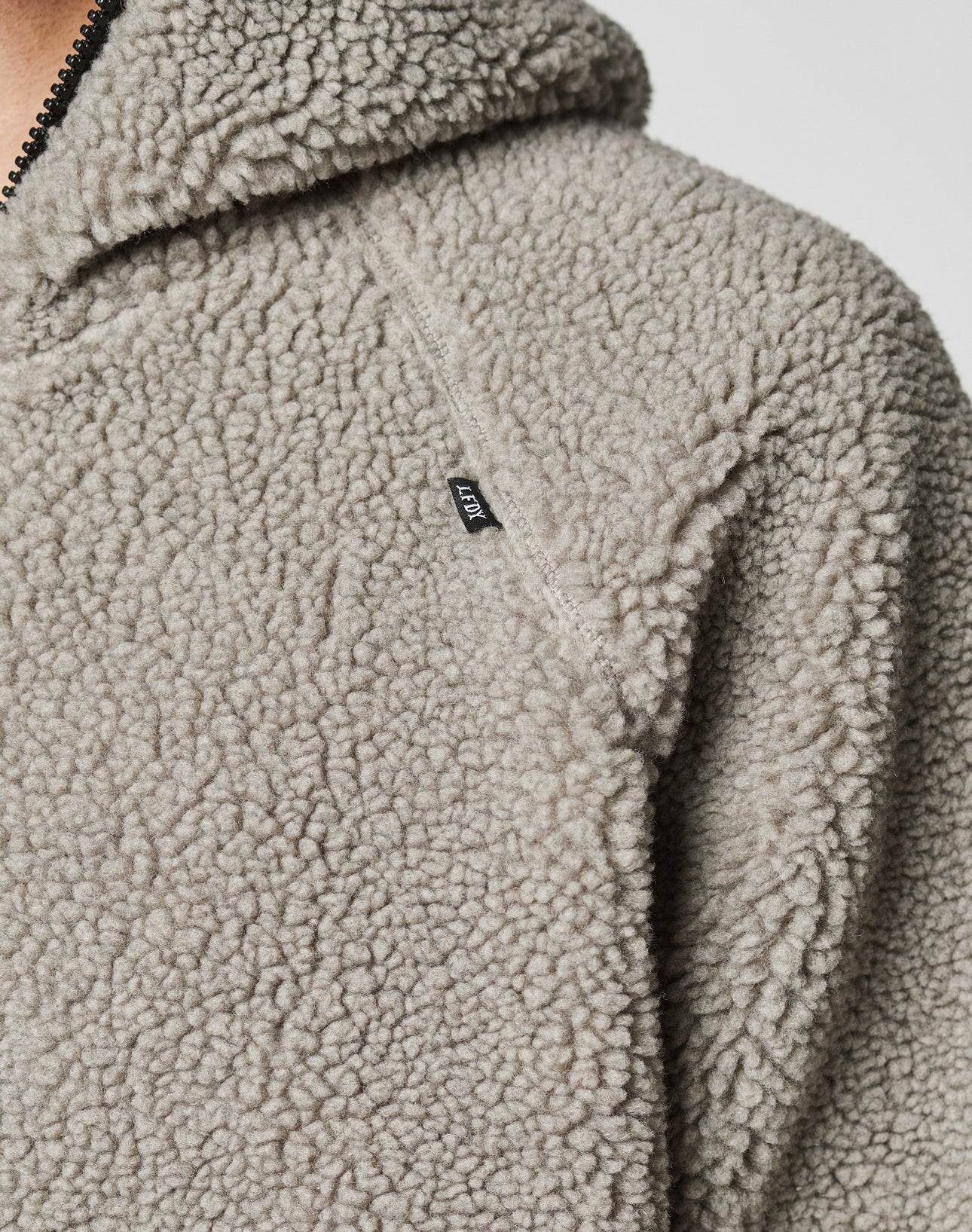 Close-up of a light gray, textured sherpa fleece Live Fast Teddy Jacket by LIVE FAST DIE YOUNG. Features a black shoulder tag, partially visible zipper, hood, and stylish nylon accents.