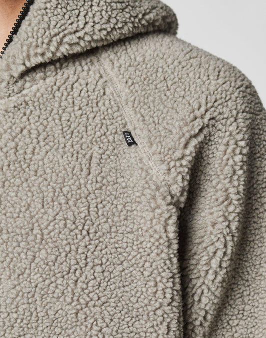 Close-up of a light gray, textured sherpa fleece Live Fast Teddy Jacket by LIVE FAST DIE YOUNG. Features a black shoulder tag, partially visible zipper, hood, and stylish nylon accents.