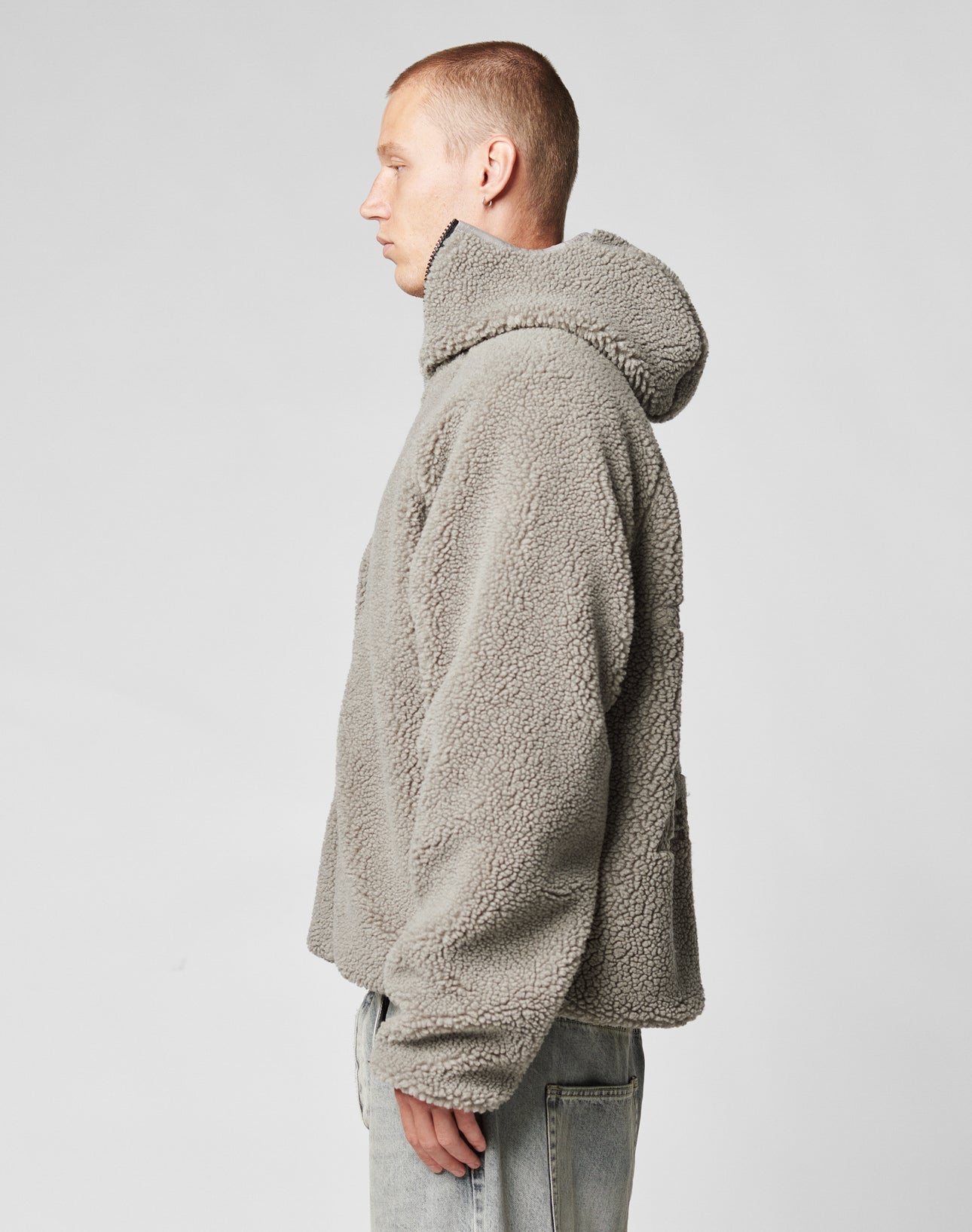 A person with short hair stands in profile against a light background, wearing an oversize fit LIVE FAST DIE YOUNG Live Fast Teddy Jacket with embossed branding and light-wash jeans.