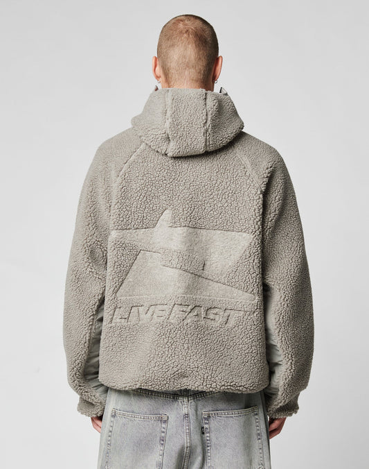 Seen from behind, a person with short hair wears the LIVE FAST DIE YOUNG Live Fast Teddy Jacket in gray fleece with a hood, featuring embossed branding and the word LIVEFAST on the back, paired with light gray jeans.