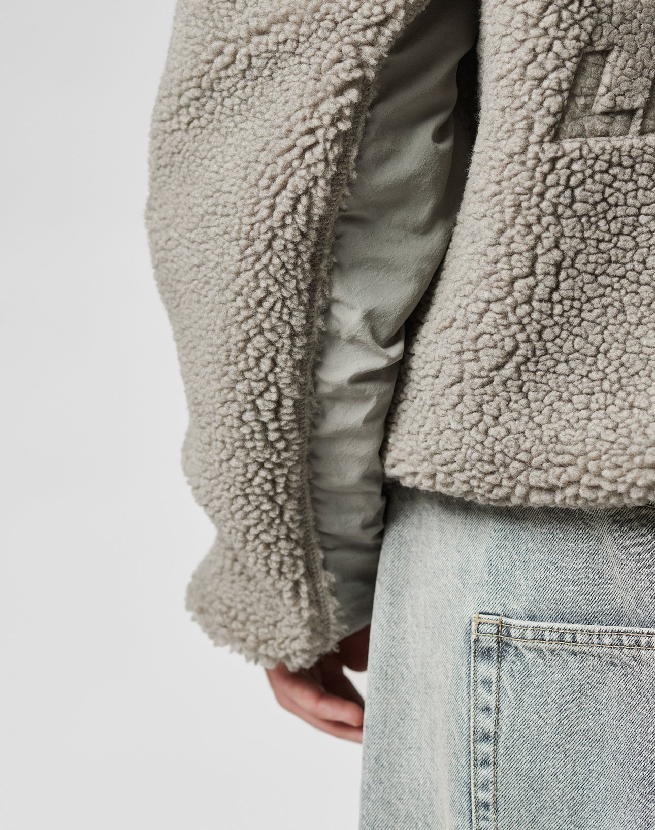 A close-up of a person wearing the LIVE FAST DIE YOUNG Live Fast Teddy Jacket with embossed branding, over a light shirt and light blue jeans. Their hand is partly visible by their side against a plain, neutral background.