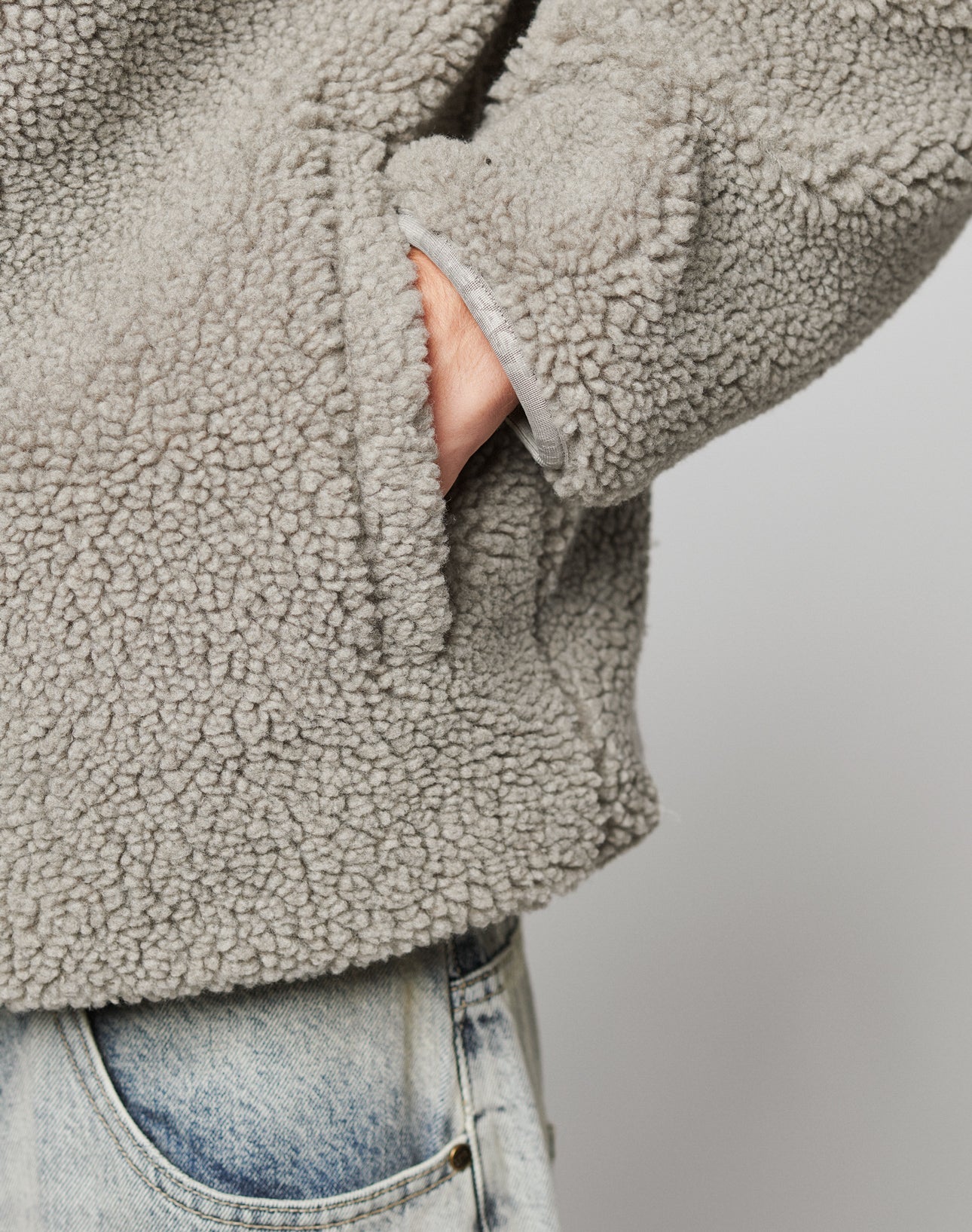 A person wearing the LIVE FAST DIE YOUNG Live Fast Teddy Jacket in light gray sherpa fleece, with one hand in the side pocket, paired with light blue jeans and subtle embossed branding, stands against a plain background.