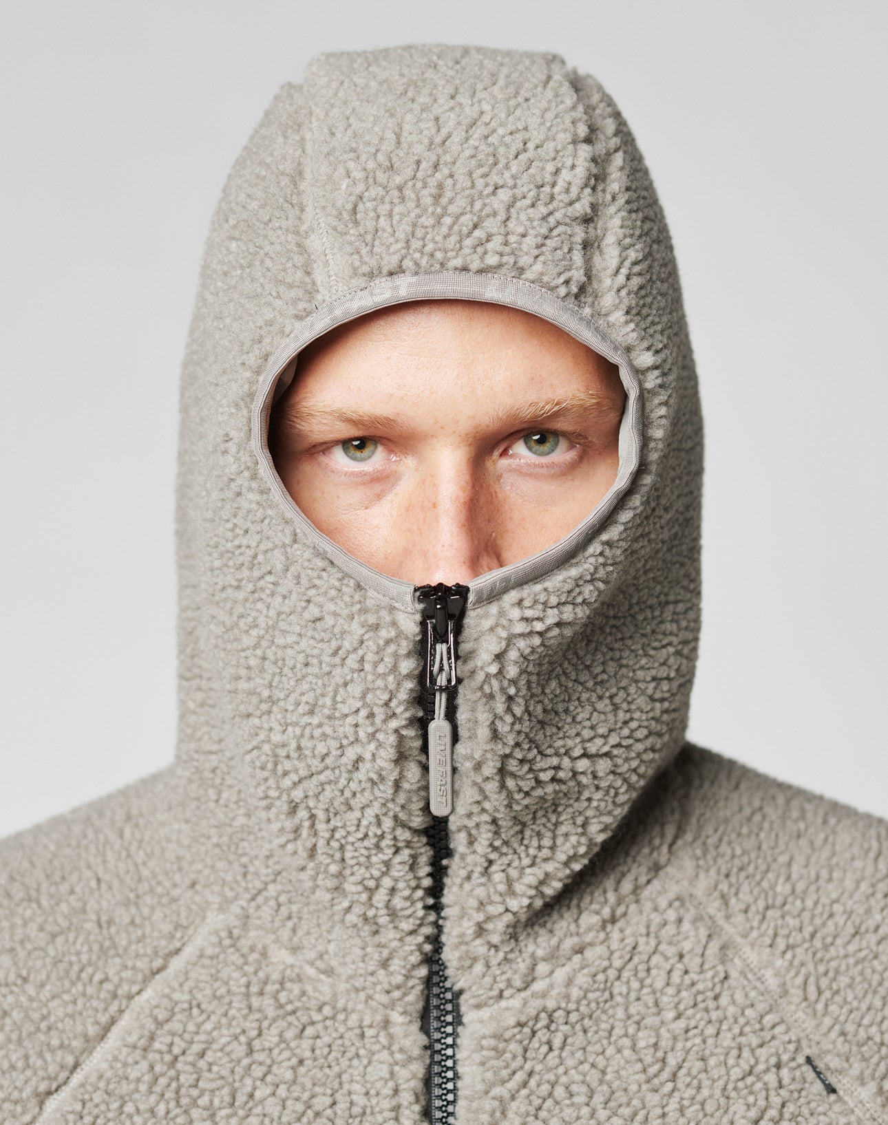 A person wears the Live Fast Teddy Jacket by LIVE FAST DIE YOUNG, zipped up to reveal only their eyes through a round opening. The high-pile fleece and subtle embossed branding stand out against the neutral background, highlighting its cozy style.