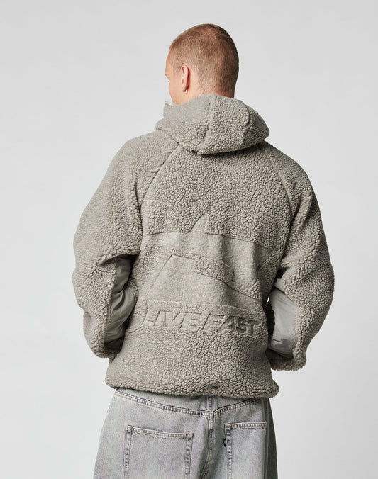 Seen from behind, a person with short hair wears the Live Fast Teddy Jacket by LIVE FAST DIE YOUNG, featuring an oversize fit, hood, and large embossed LIVE FAST branding on the back. They style it casually with light-wash jeans.