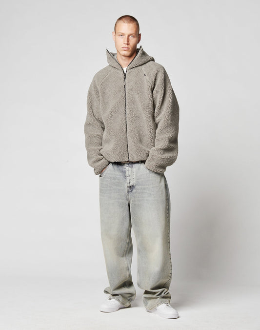A person with short hair wears the LIVE FAST DIE YOUNG Live Fast Teddy Jacket, featuring a gray zip-up fleece and hood, loose faded jeans, and white sneakers, standing against a plain light gray background.