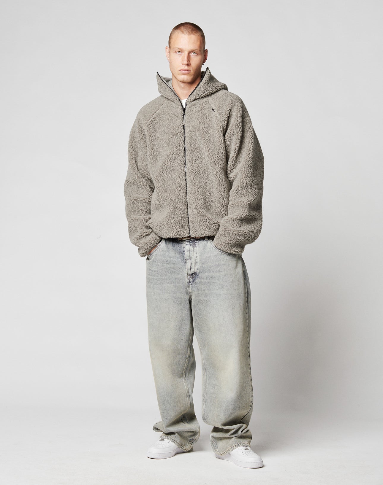 A person with short hair wears the LIVE FAST DIE YOUNG Live Fast Teddy Jacket, featuring a gray zip-up fleece and hood, loose faded jeans, and white sneakers, standing against a plain light gray background.