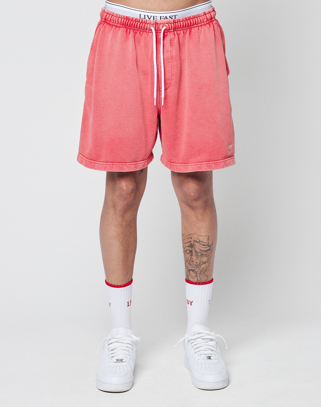 A person wears LIVE FAST DIE YOUNG Basic Sweat Shorts in coral pink with an elastic waistband and handcrafted fade effect, white socks with red accents, and white sneakers. Only their lower body is visible against a plain light background.