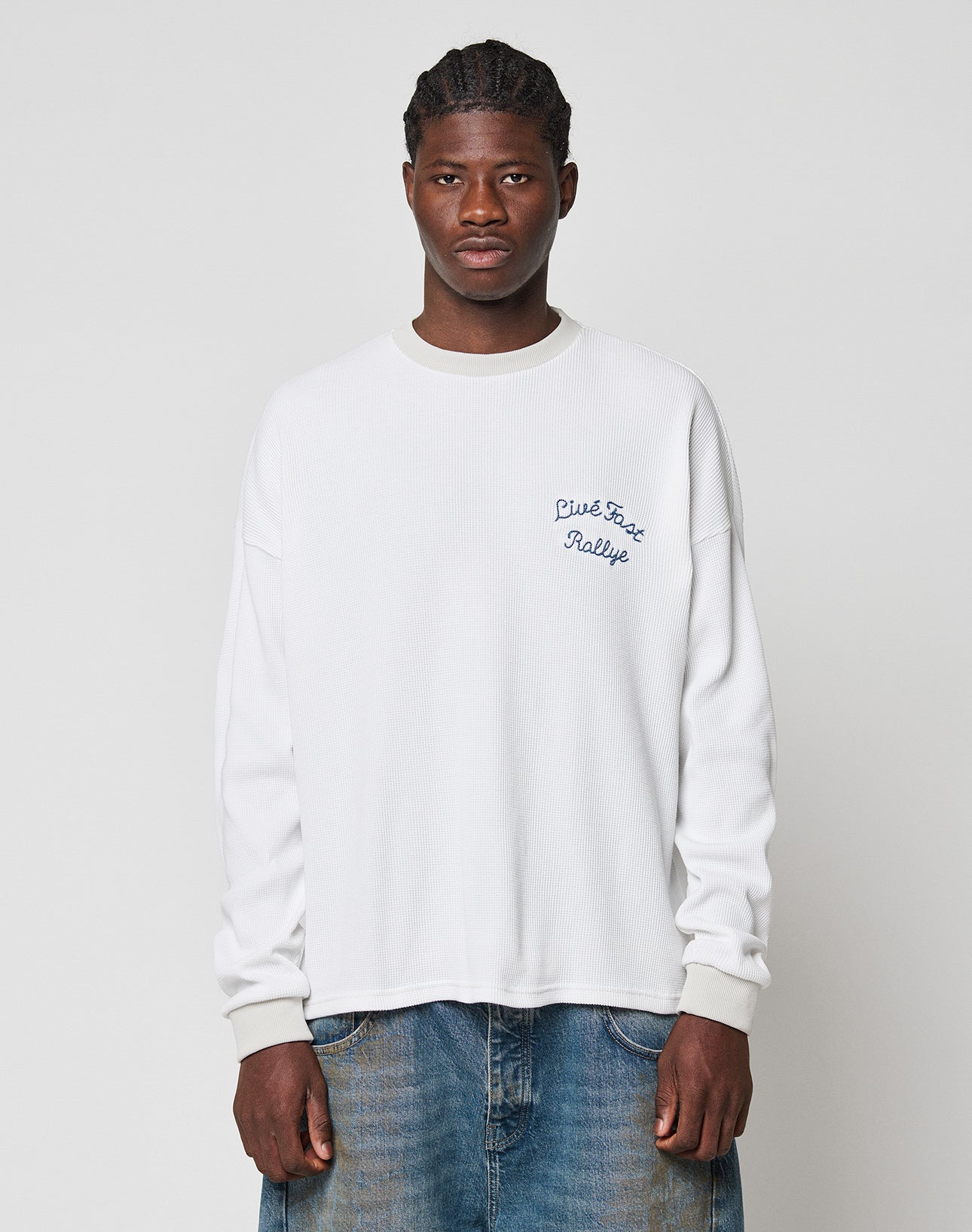 A man with short braided hair and a neutral expression wears the LIVE FAST DIE YOUNG LÉquipe Waffle Longsleeve—an oversized 100% Baumwolle white shirt with “C’est Facile Rallye” on the chest, subtle backprint detail, and blue jeans.