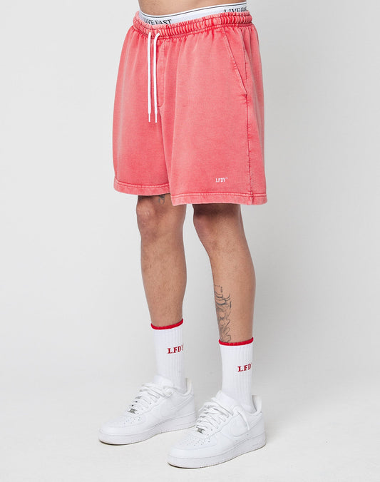 A person wears LIVE FAST DIE YOUNG Basic Sweat Shorts in coral pink with a handmade fade effect, white crew socks with red accents, and white sneakers. Only the lower half of the body is shown against a plain white background.