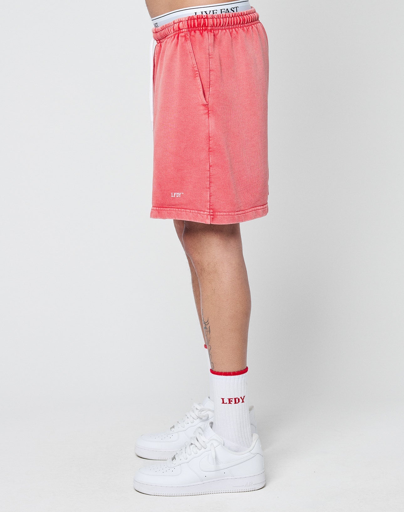 A person stands sideways wearing LIVE FAST DIE YOUNG Basic Sweat Shorts in pink, white “LFDY” socks, and white sneakers. Only the lower half of their body is shown against a plain white background.