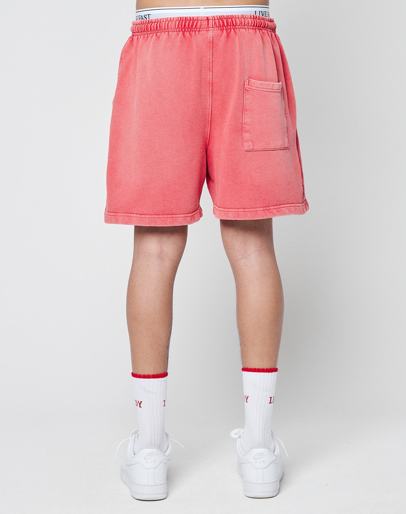 A person faces away, wearing LIVE FAST DIE YOUNG Basic Sweat Shorts in coral pink with elastic waistband and handmade fade effect, plus white socks with red trim and white sneakers, set against a plain light background.