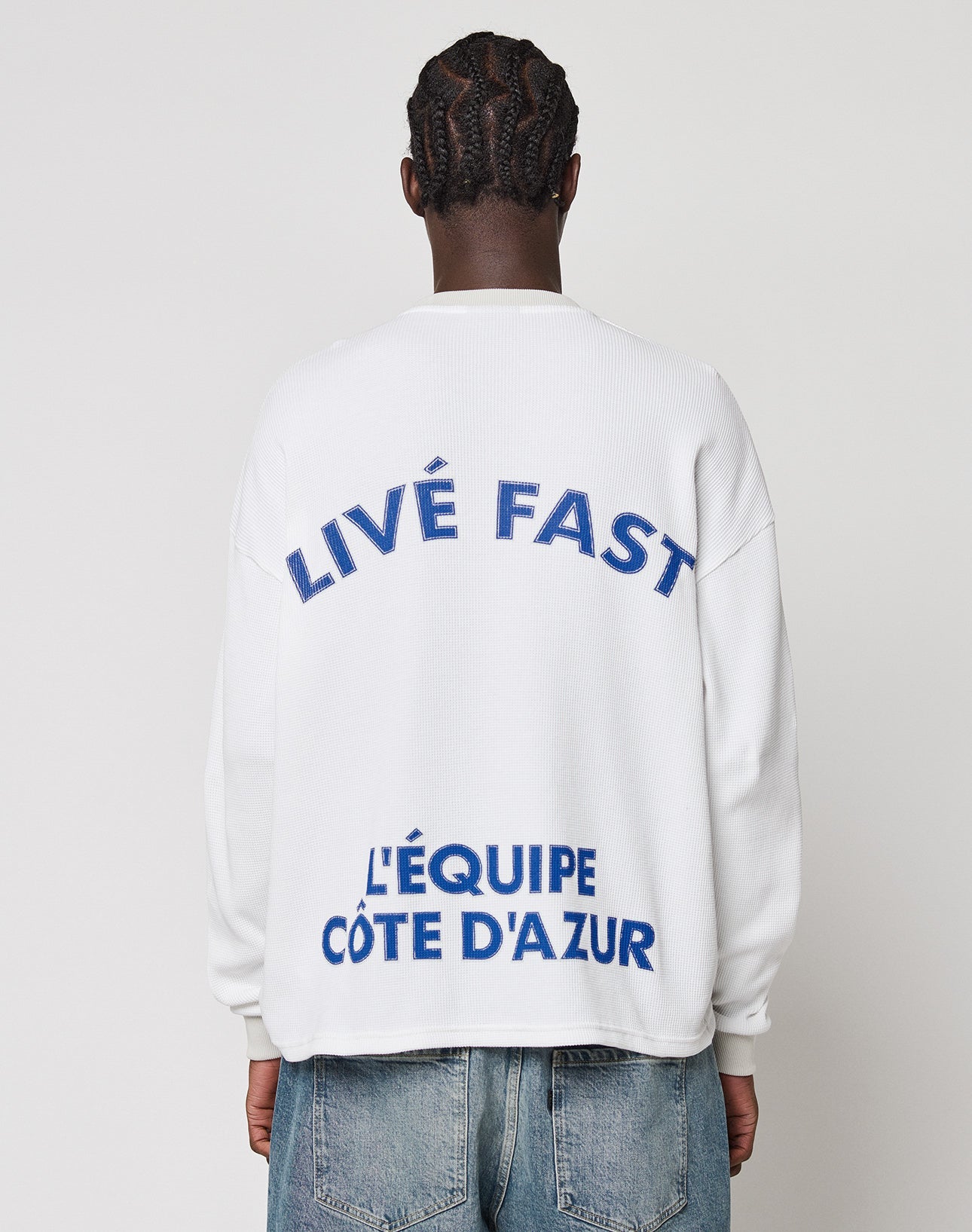 Person seen from behind in the LÉquipe Waffle Longsleeve by LIVE FAST DIE YOUNG, an oversize-fit white sweatshirt with blue LIVE FAST and LÉQUIPE CÔTE DAZUR backprint, paired with blue jeans. Made from 100% Baumwolle. White background.