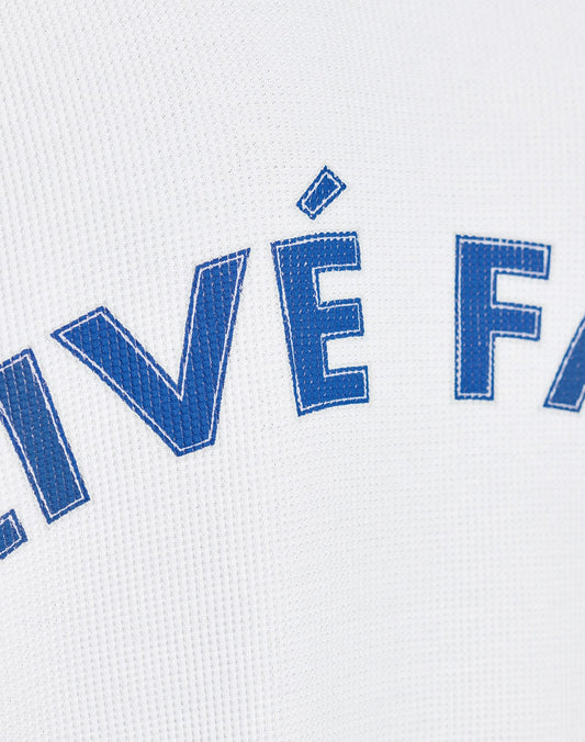 Close-up of blue letters with white stitching on textured white fabric, partially showing VÉ FA. The LÉquipe Waffle Longsleeve by LIVE FAST DIE YOUNG features 100% Baumwolle and an Oversize Fit for extra comfort.