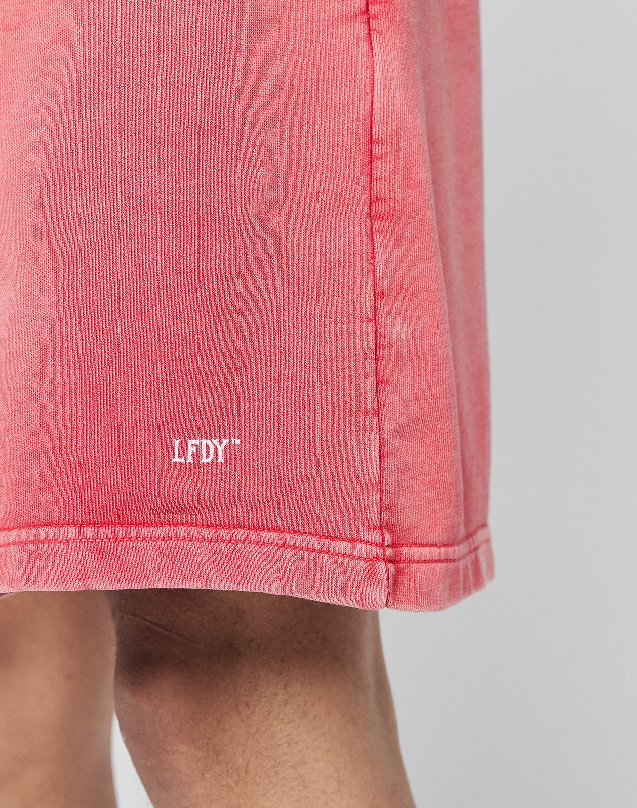 A close-up of a person wearing LIVE FAST DIE YOUNG Basic Sweat Shorts in pink, featuring an elastic waistband and a white LFDY™ logo near the hem. Only part of the leg and the subtly faded lower shorts are visible.