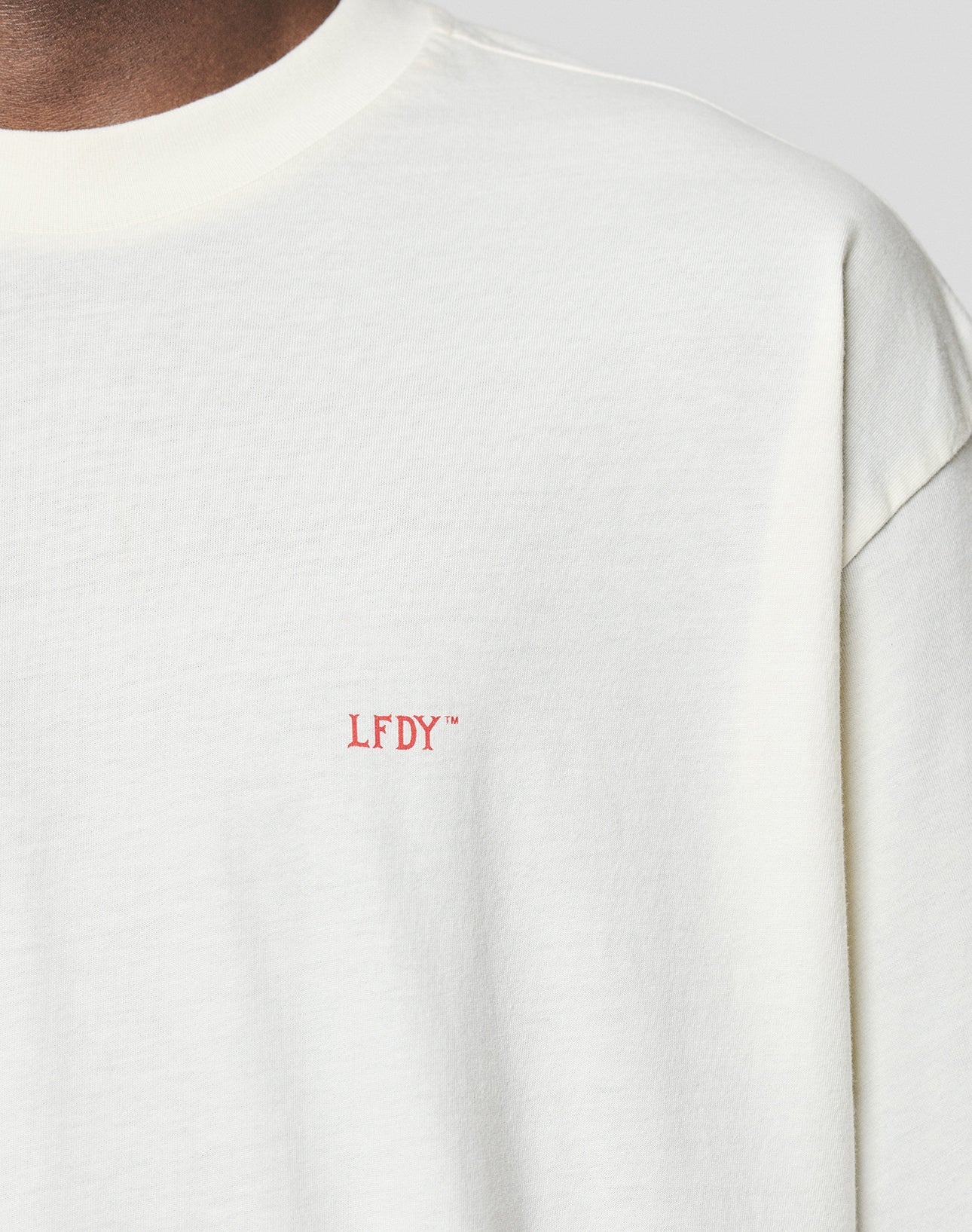 A close-up shows someone wearing the LIVE FAST DIE YOUNG LFDY Flash Tee, an off-white oversized shirt with red LFDY embroidery on the chest, highlighting its premium 160gsm fabric. Only part of the neck and shoulder is visible.