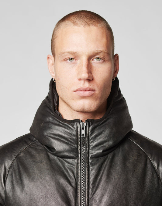 A person with short hair and light skin models the LIVE FAST DIE YOUNG Live Fast Leather Puffer Jacket—black Schafsleder with a high padded collar, oversize fit, and embossed Live Fast Logo—looking neutrally at the camera against a plain background.