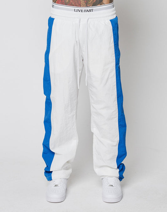 A person models the LIVE FAST DIE YOUNG LFDY Trackpants 3000—wide fit, white with blue side panels, and an elastic waistband reading LIVE FAST—paired with white sneakers. Photo shows waist down on a plain white background.