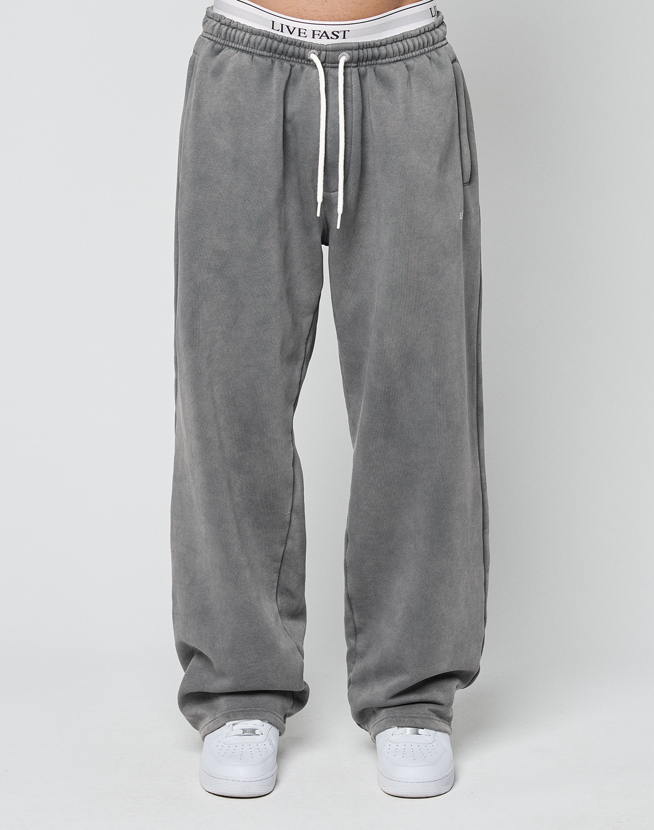 A person wears the LIVE FAST DIE YOUNG Basic Wide Jogger—oversized fit gray sweatpants with white drawstrings and white sneakers, standing against a plain light background. The waistband showcases the LIVE FAST logo for a bold accent.
