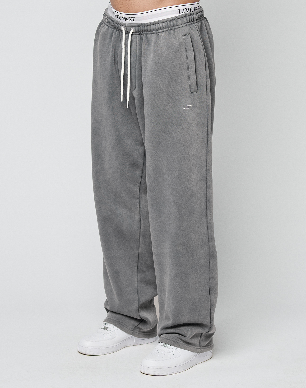 A person wearing LIVE FAST DIE YOUNG Basic Wide Jogger sweatpants in gray with a left thigh logo, wide fit, and white sneakers stands against a plain white background. | model