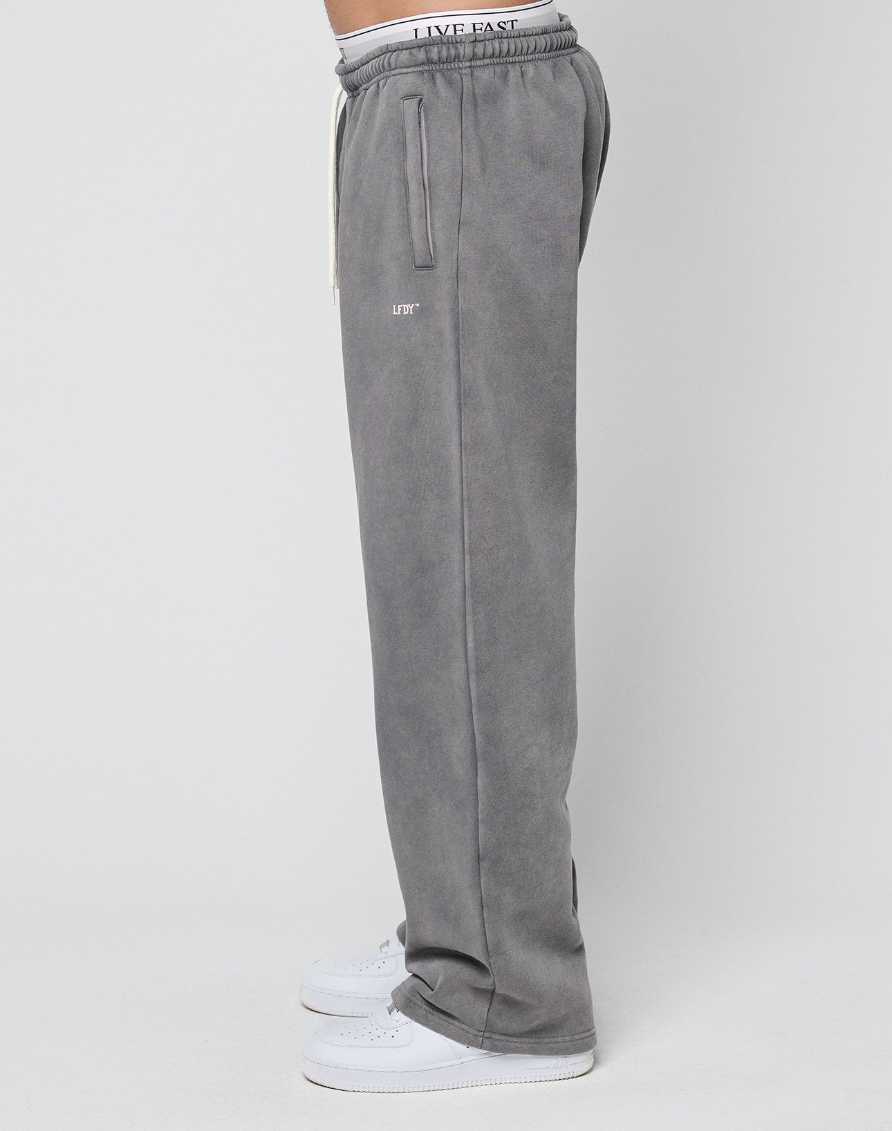 A person stands sideways in Basic Wide Jogger sweatpants by LIVE FAST DIE YOUNG, featuring an oversize fit, vertical pockets, and a visible elastic waistband. They pair them with white sneakers against a plain light background.