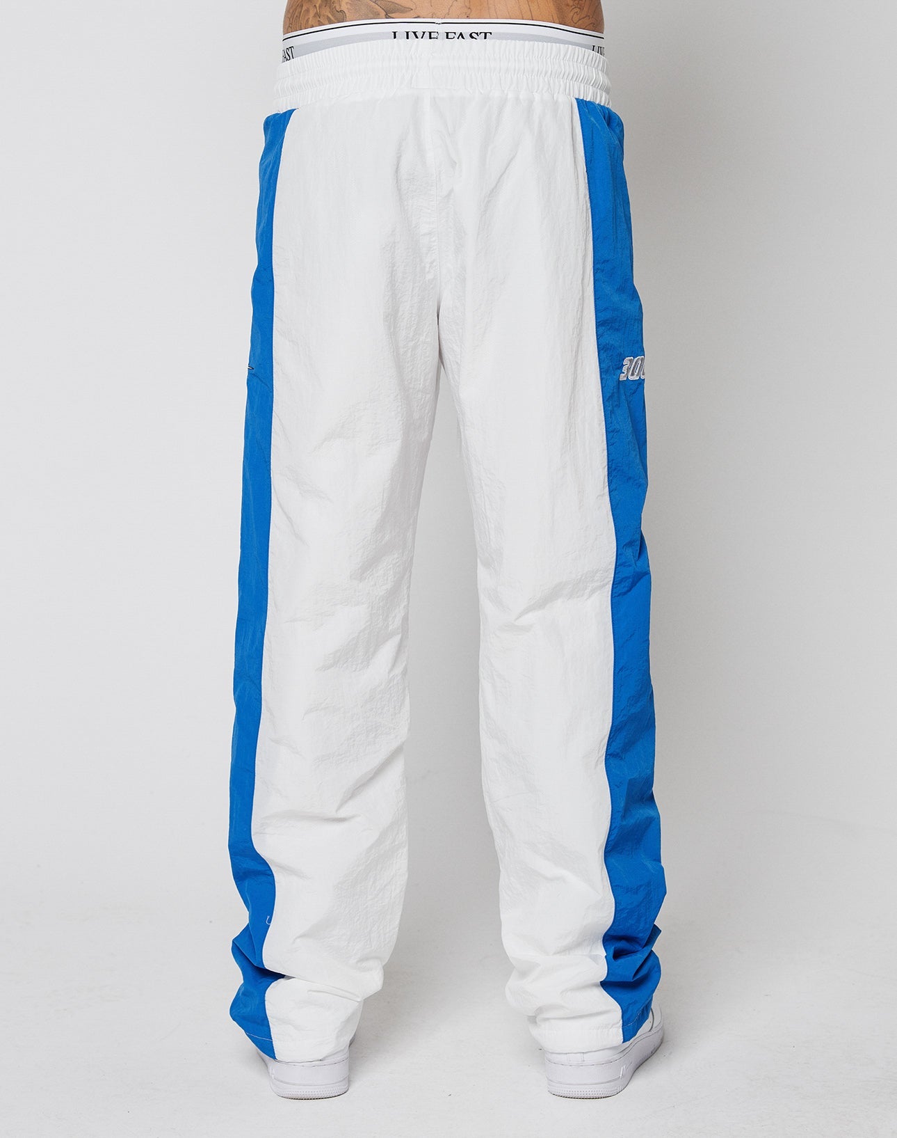 The lower half of a person is shown wearing LIVE FAST DIE YOUNG LFDY Trackpants 3000, featuring wide fit, white color with blue side panels and elastic waistband, paired with white shoes against a plain white background.