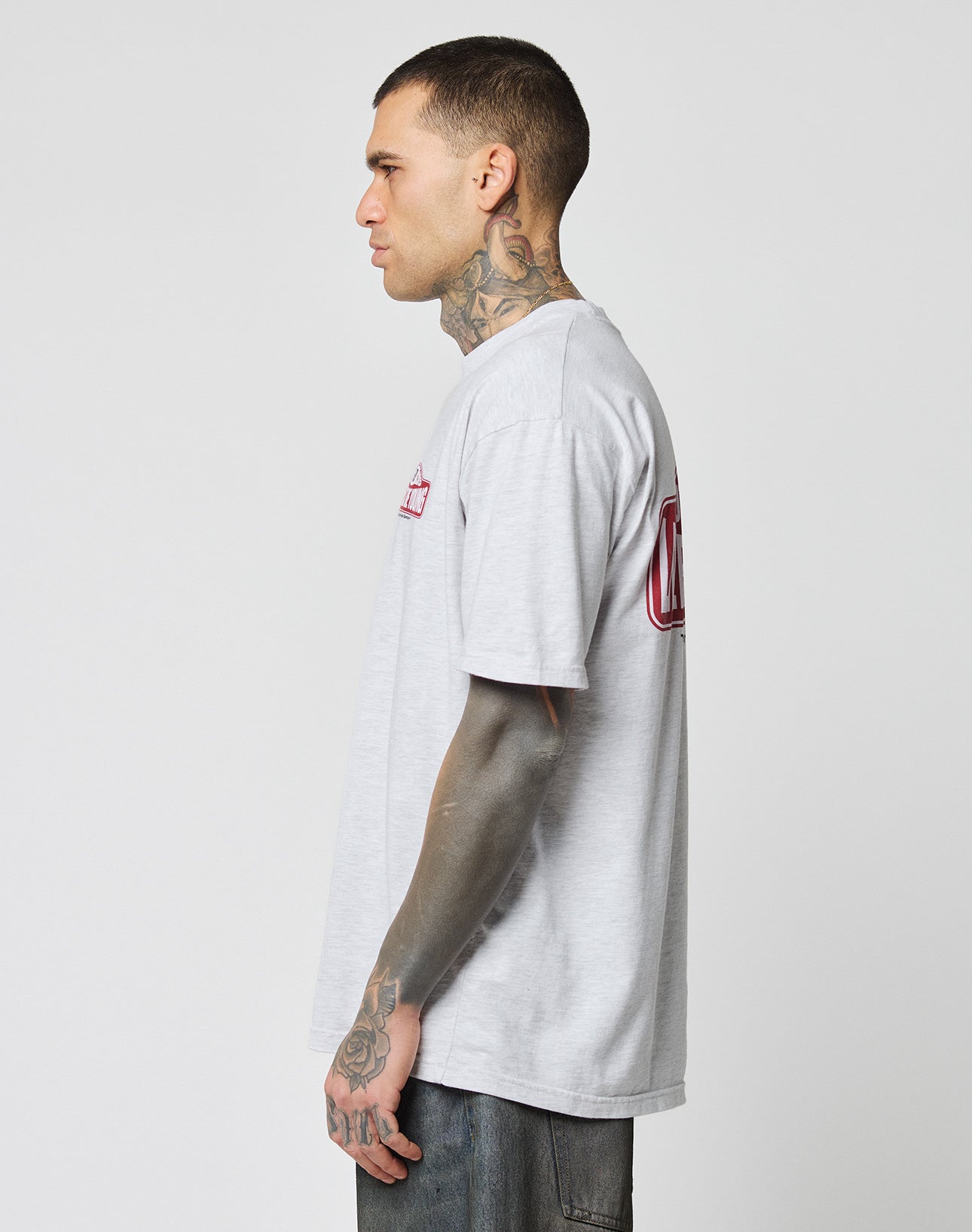 A tattooed man stands in profile wearing the LIVE FAST DIE YOUNG Live Fast 2013 Tee, a Regular Fit light gray 100% Baumwolle T-shirt, and dark jeans against a plain white background.