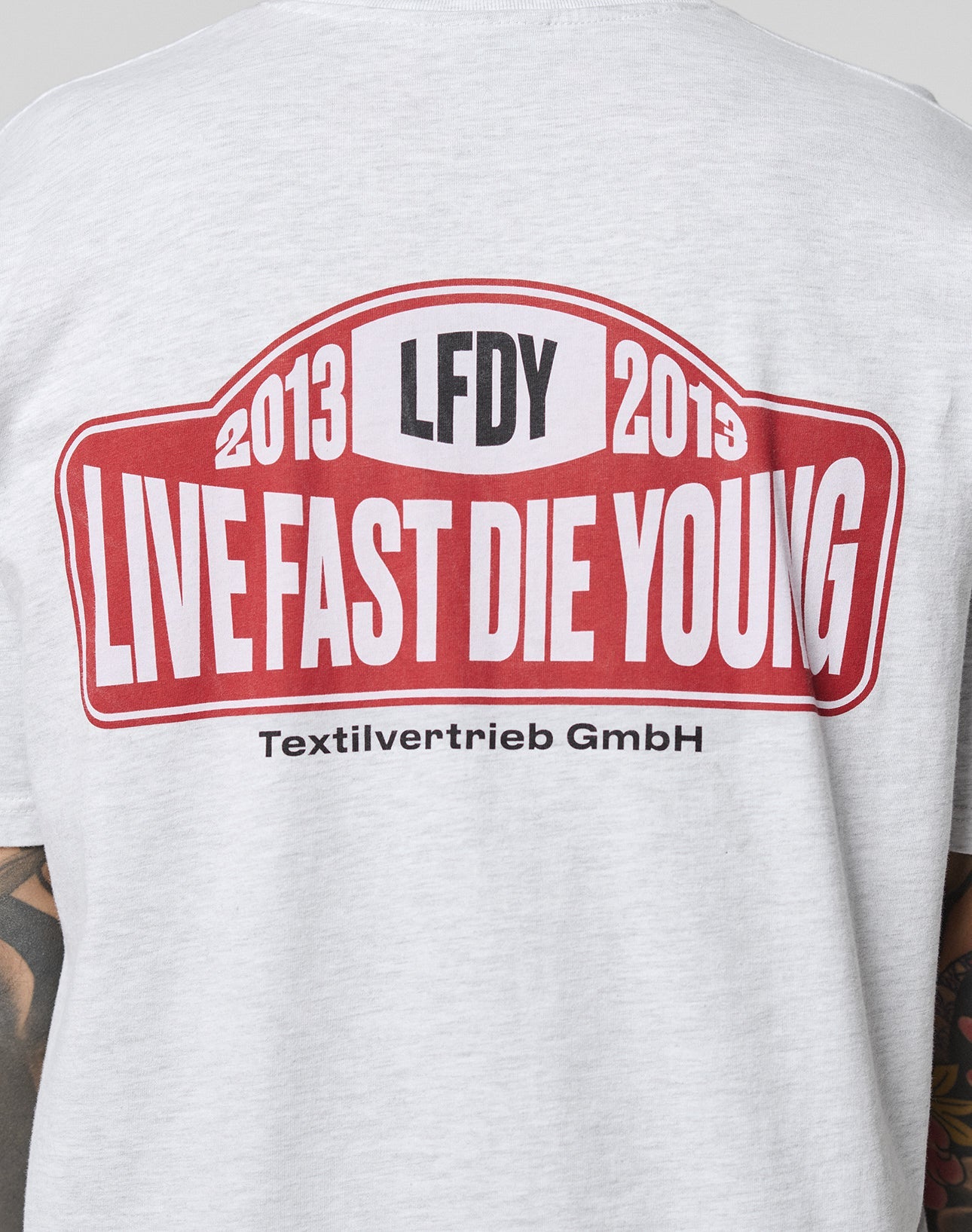 A person wears the Live Fast 2013 Tee by LIVE FAST DIE YOUNG—a light gray, regular fit T-shirt with a bold red and white backprint. The tee is made from 100% Baumwolle and partially reveals tattooed arms.