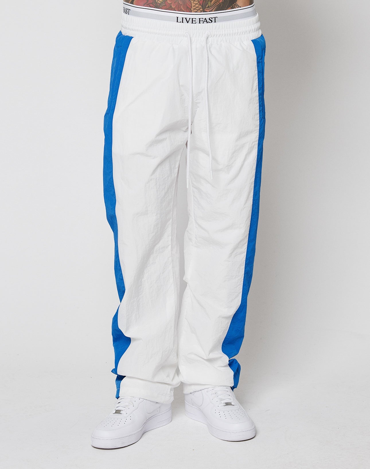 A person wears LIVE FAST DIE YOUNGs LFDY Trackpants 3000—wide fit, white with bright blue side stripes and a bold LIVE FAST elastic waistband—paired with white sneakers, shown from the waist down.