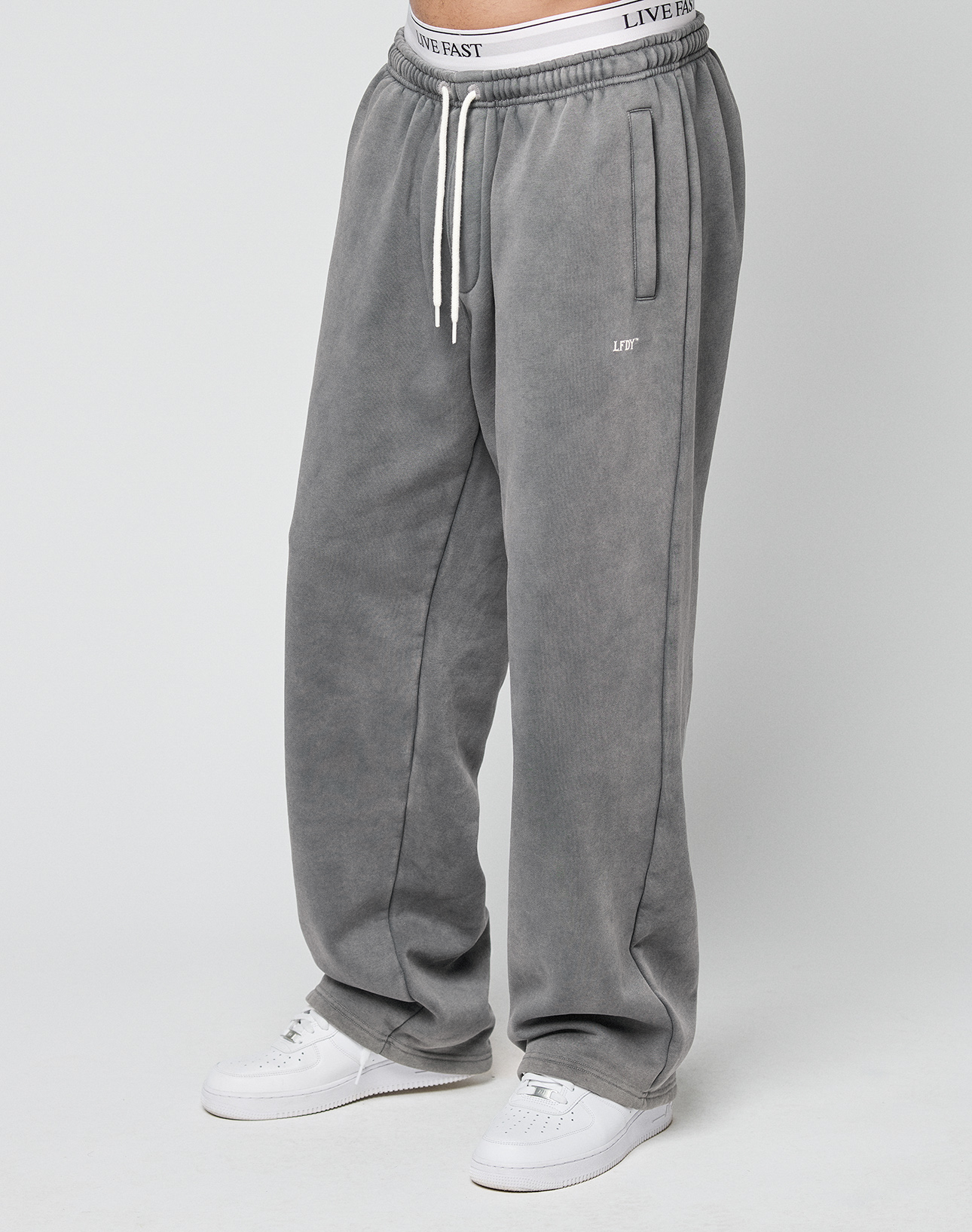Person wearing LIVE FAST DIE YOUNG Basic Wide Jogger in gray with an oversized fit and white drawstrings, paired with white sneakers. Only the body from the waist down is visible. LIVE FAST logo is printed on the waistband.
