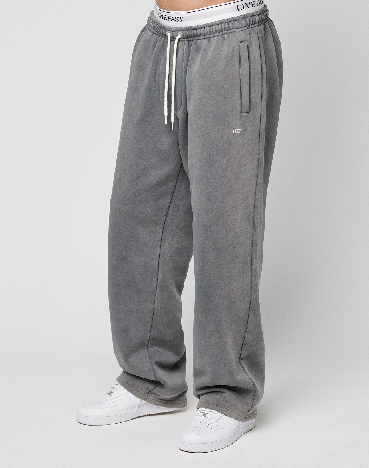 A person wears the LIVE FAST DIE YOUNG Basic Wide Jogger in Oversize Fit—loose gray sweatpants with white drawstrings and an elastic waistband showing LIVE FAST text—paired with white sneakers against a plain light gray background.
