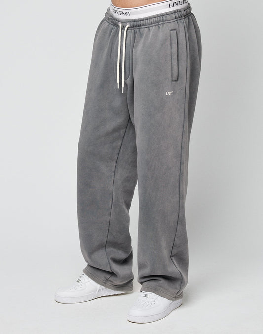 A person wears the LIVE FAST DIE YOUNG Basic Wide Jogger in Oversize Fit—loose gray sweatpants with white drawstrings and an elastic waistband showing LIVE FAST text—paired with white sneakers against a plain light gray background.