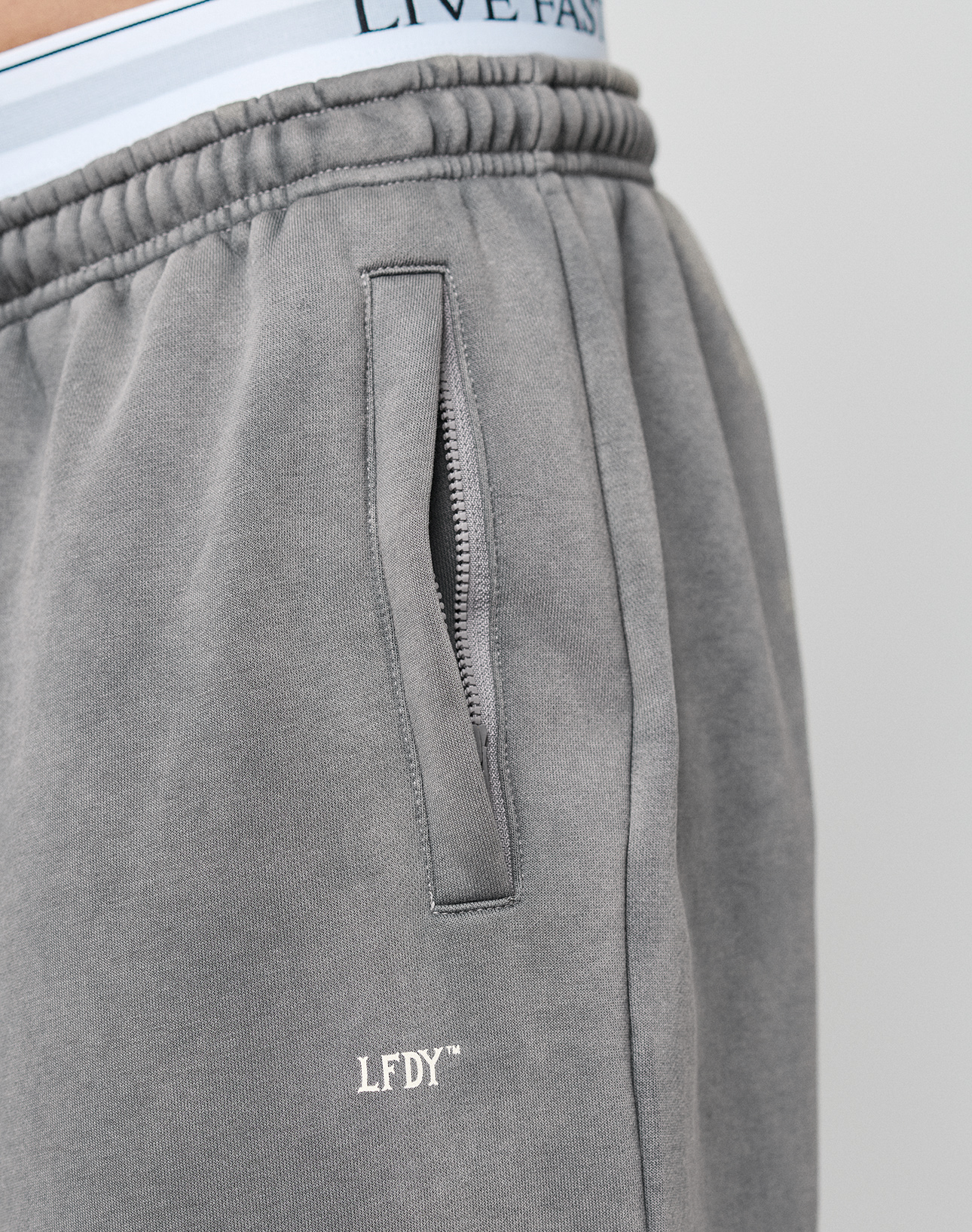 Close-up of a person wearing the LIVE FAST DIE YOUNG Basic Wide Jogger in grey with oversized fit, zippered pocket, LFDY logo print, and light blue underwear featuring LIVE FAST text visible above the waistband.