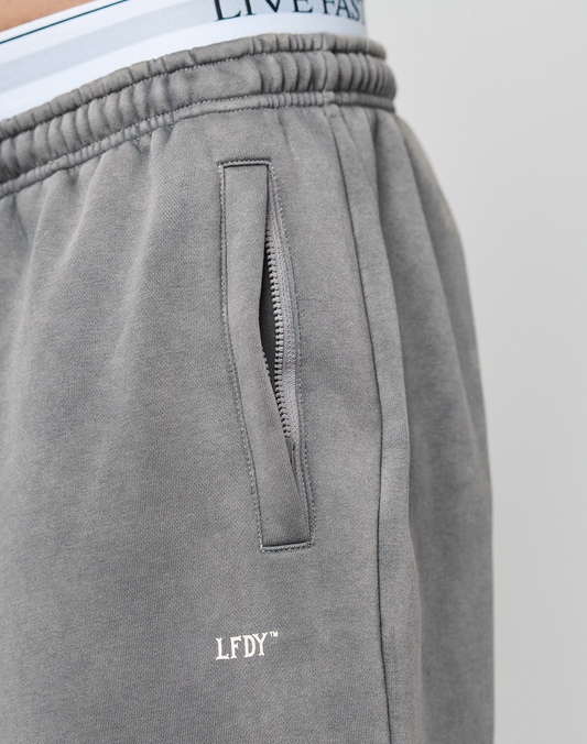 Close-up of a person wearing the LIVE FAST DIE YOUNG Basic Wide Jogger in grey with oversized fit, zippered pocket, LFDY logo print, and light blue underwear featuring LIVE FAST text visible above the waistband.