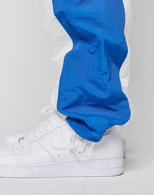 A person wears LIVE FAST DIE YOUNG LFDY Trackpants 3000 in bright blue and white with an elastic waistband and cuffs, paired with clean white Nike Air Force 1 sneakers, standing on a white surface against a plain white background.
