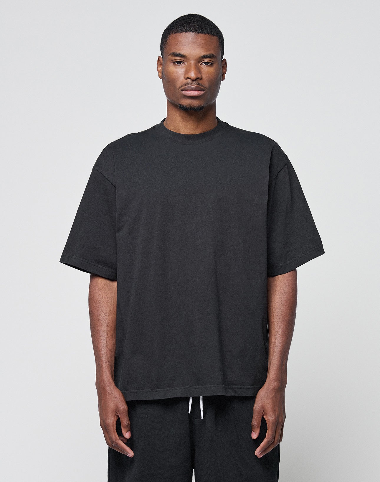 A man faces forward against a plain light gray background, wearing the LIVE FAST DIE YOUNG Basic Heavy Tee in oversize black 100% Baumwolle and black pants, with a neutral expression and arms relaxed at his sides. | model