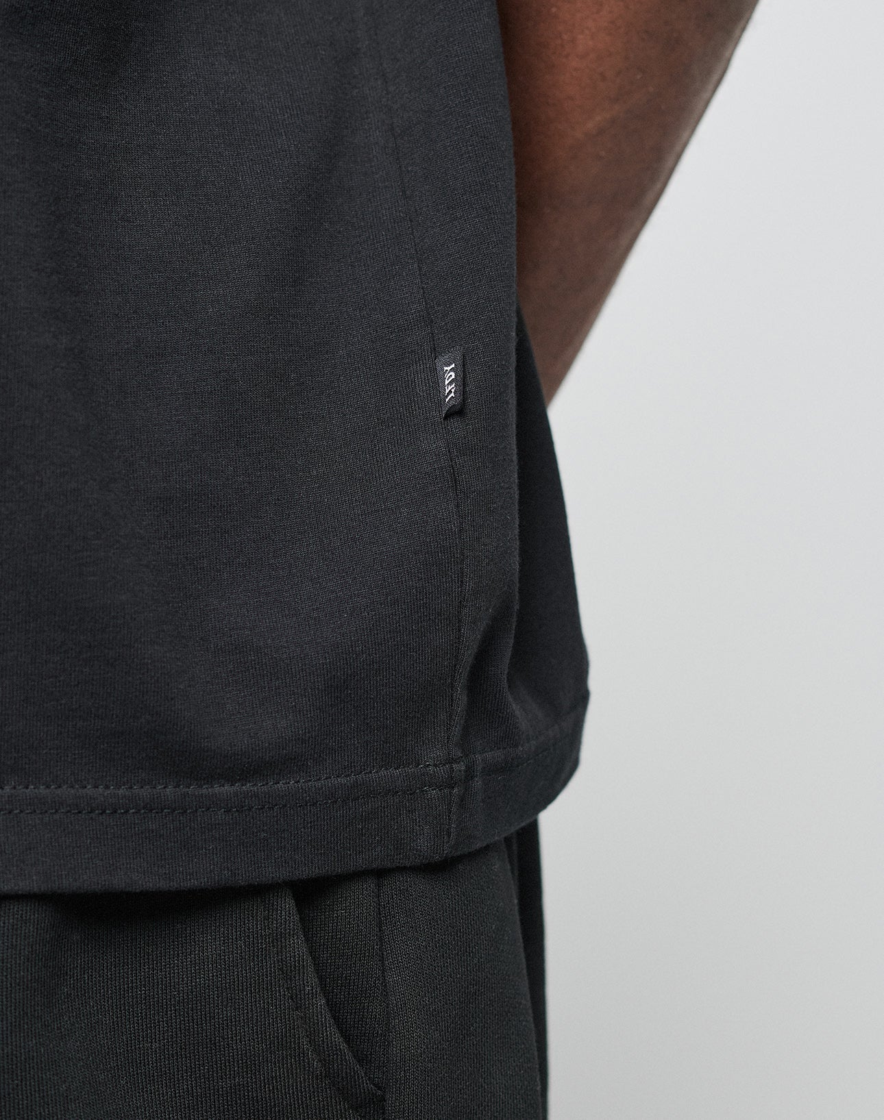 Close-up of a person wearing the LIVE FAST DIE YOUNG Basic Heavy Tee in black, styled with black pants. A small black tag with white text is sewn into the tee’s side seam. Only the lower torso and part of the arm are visible.