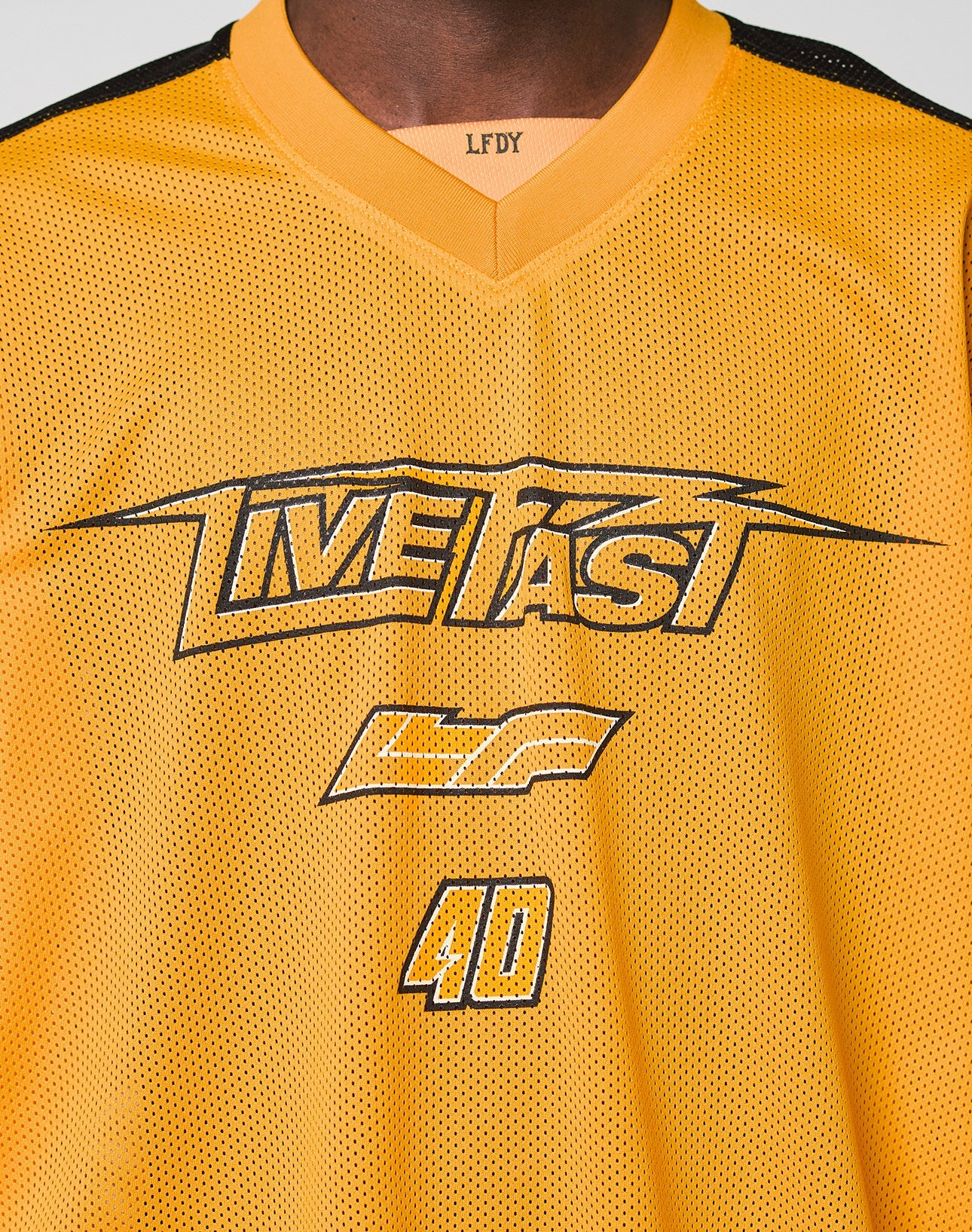 A close-up of a person wearing the LFDY Autobahn Jersey by LIVE FAST DIE YOUNG, featuring a yellow mesh polyester fabric, oversize fit, black “LIVE FAST” and “GF 40” prints on the front, and multilogo details including LFDY on the neckline.