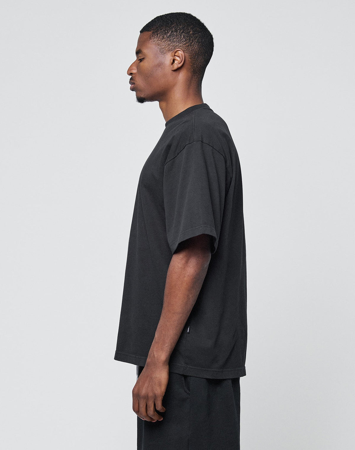 A man stands in profile against a light background, wearing the LIVE FAST DIE YOUNG Basic Heavy Tee—an oversized black T-shirt made of 100% cotton—and black pants. He looks left, arms relaxed at his sides. Please follow care instructions.