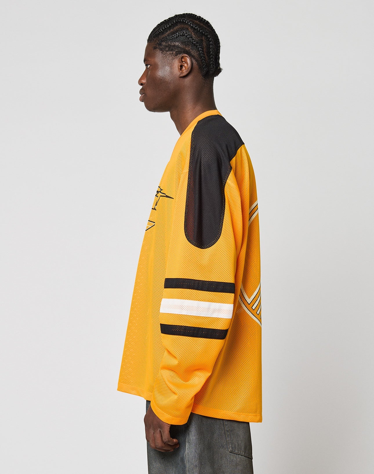 A person with braided hair stands in profile wearing the LIVE FAST DIE YOUNG LFDY Autobahn Jersey—an oversized yellow sports top with black and white striped sleeves and black shoulder patch—paired with gray pants against a plain background.