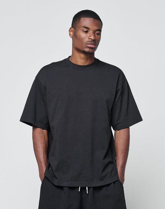 A man with short hair wears the LIVE FAST DIE YOUNG Basic Heavy Tee, a loose-fitting black 100% cotton T-shirt, and black pants, standing against a plain light gray background with his hands in his pockets, looking slightly downward.