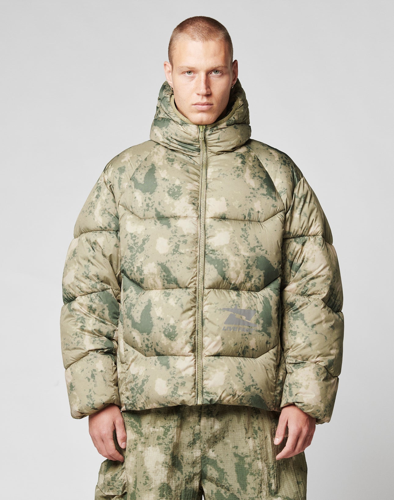 A person with short hair wears the LIVE FAST DIE YOUNG Live Fast Puffer Jacket—an oversize green and beige camo style with a hood and reflective front and back print—posing against a plain light gray background.