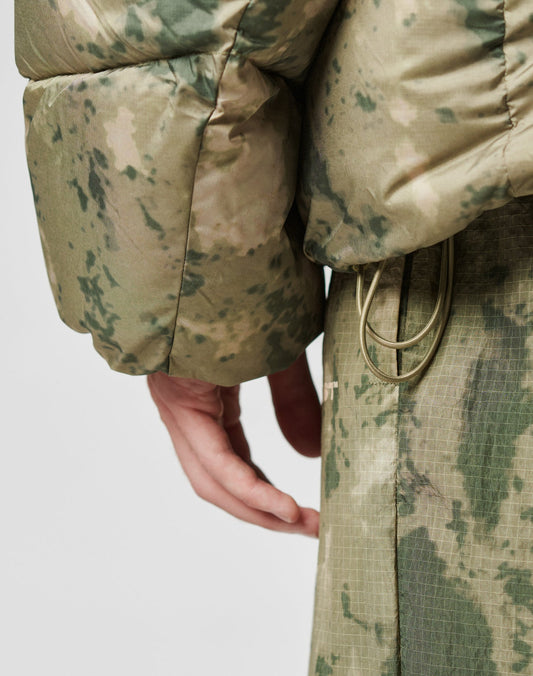Close-up of a persons hand by their side, wearing the LIVE FAST DIE YOUNG Live Fast Puffer Jacket in green and beige camouflage with an oversized fit and drawstrings at the waist, paired with matching pants.