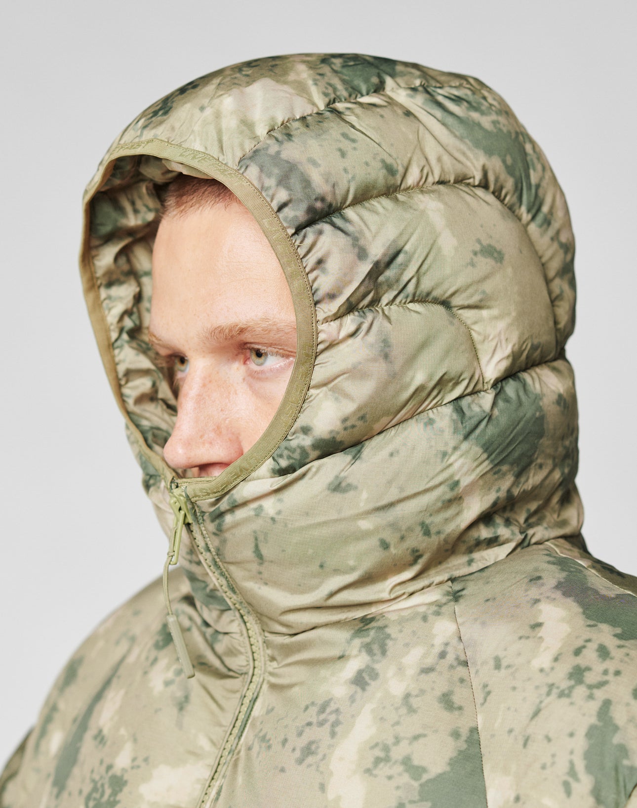 Someone wears the LIVE FAST DIE YOUNG Live Fast Puffer Jacket in green camouflage, with an Oversize Fit and the hood up, covering most of their head and framing their face in a side profile.