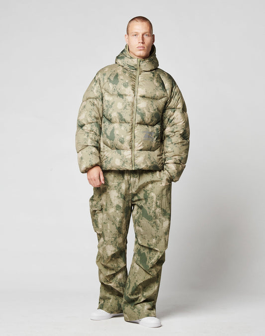 A person stands facing forward, wearing the LIVE FAST DIE YOUNG Live Fast Puffer Jacket in green and tan camouflage with matching cargo pants and white sneakers, set against a plain light gray backdrop.