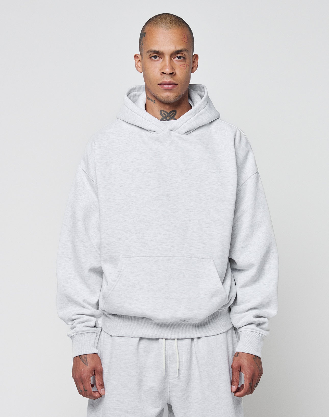 A person with a shaved head models the LIVE FAST DIE YOUNG Basic Blank Hooded and matching sweatpants in light gray, featuring an oversize fit and soft Baumwolle fabric, standing forward against a plain background.