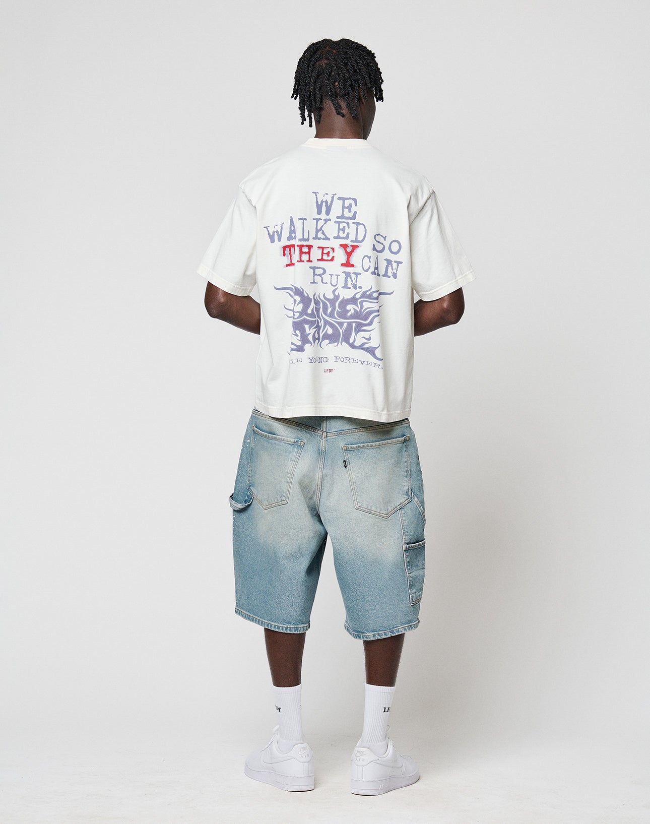 A person faces away wearing the LIVE FAST DIE YOUNG LFDY We Walked Tee—an oversized white graphic T-shirt with purple and red front and back text—paired with light blue denim shorts, white socks, and sneakers against a plain white background.