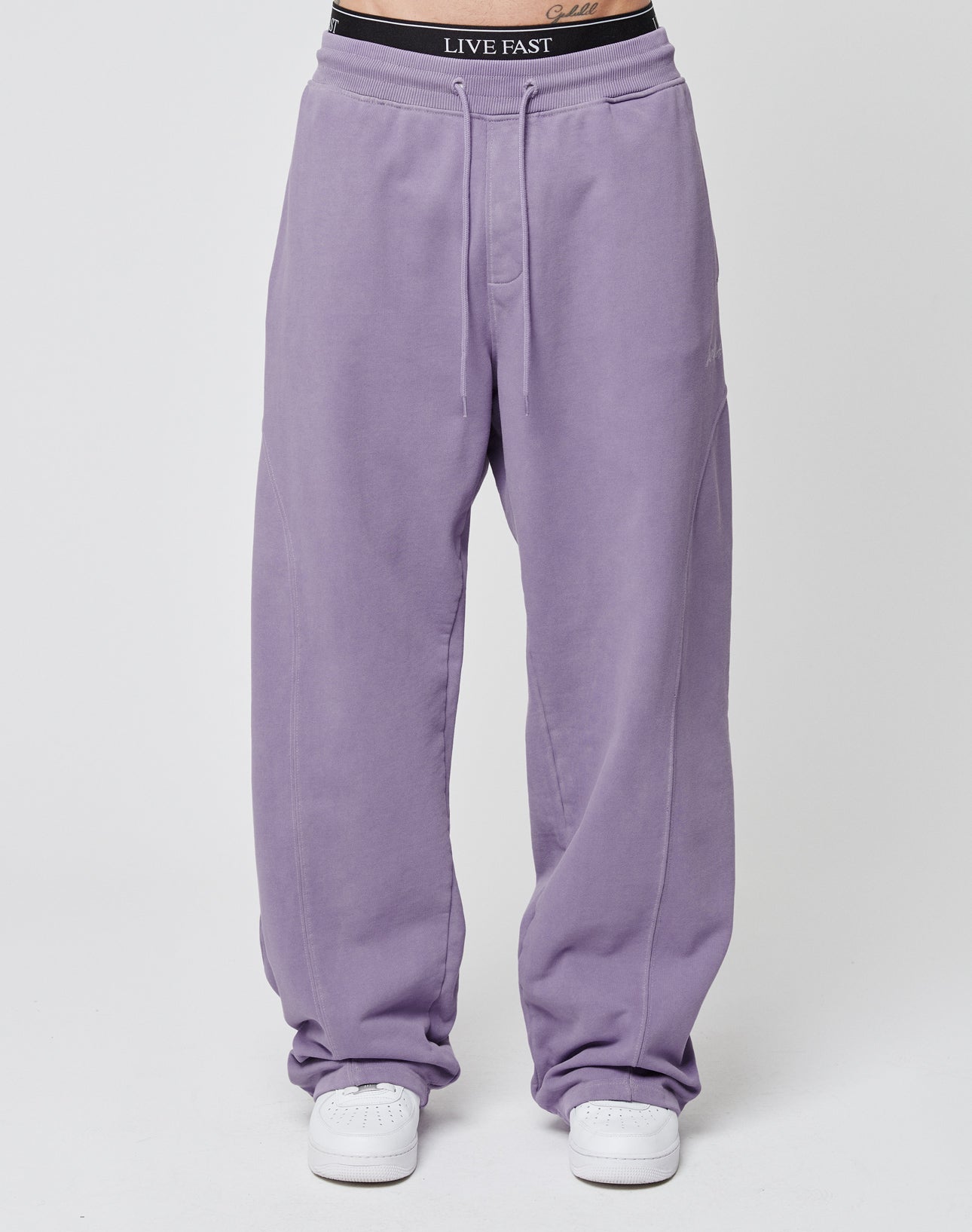 A person models the LIVE FAST DIE YOUNG Basic Wide Jogger in lilac with an elastic waistband, paired with white sneakers and black underwear featuring a LIVE FAST waistband, standing against a plain white background.