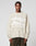 A man with short curly hair models the LIVE FAST DIE YOUNG Live Fast Fuzzy Sweater—an oversized, light beige polyamide-elastane blend with a white abstract intarsia logo and “LIVE FAST” on the front—paired with dark pants against a gray background.