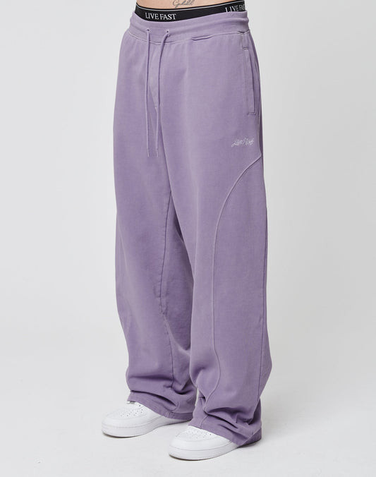 A person wears the Basic Wide Jogger by LIVE FAST DIE YOUNG in lavender, made from soft cotton with a wide fit, subtle seam details, and a black elastic waistband that says LIVE FAST. Styled with white sneakers on a plain white background.